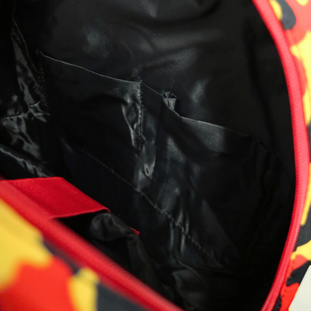 Close-up of the interior of a bag or backpack, showing a black satin-like lining with a side pocket and a red zipper edge. The outer fabric has a partial red, yellow, and black pattern visible at the edges.