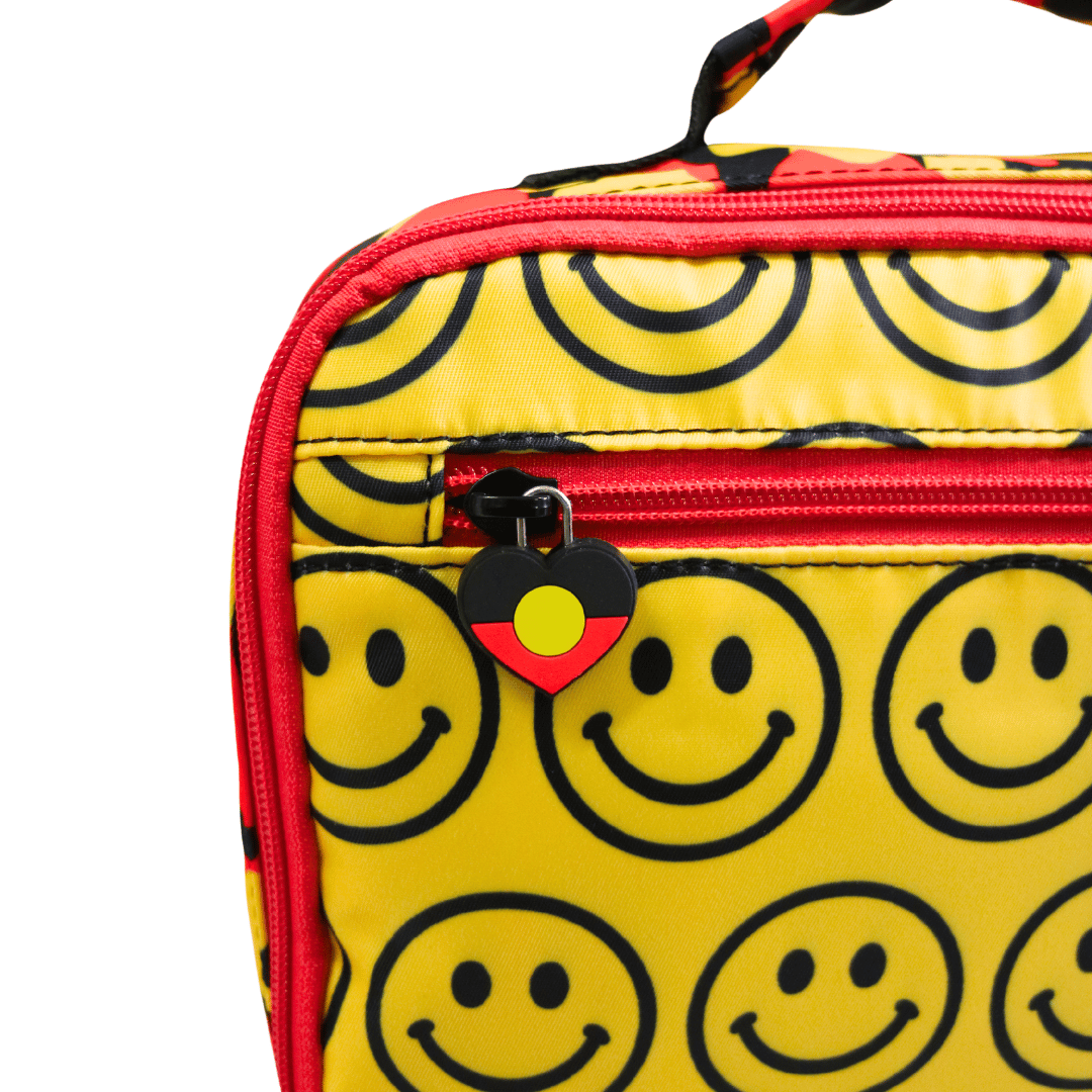 Close-up of haus of ziggy lee everyday deadly yellow lunch bag with a black smiley face pattern, red zipper trim, and a custom aboriginal flag heart-shaped zipper pull